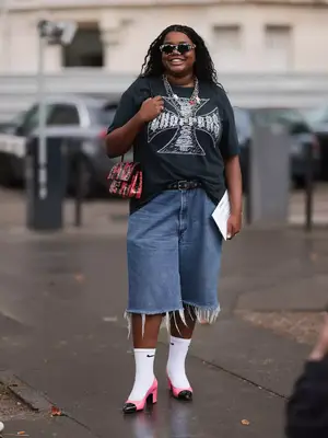 Gabriella Karefa-Johnson wears a black band tee, patterned sunglasses, red and black Chanel bag, long denim shorts, Nike socks, and two-toned patent heels