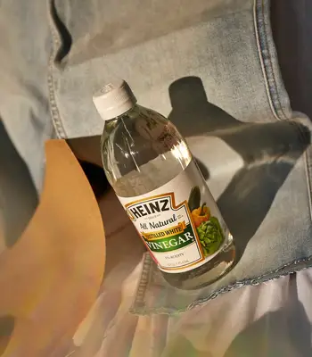 Bottle of distilled white vinegar sitting on top of pair of light wash jeans with sandpaper nearby