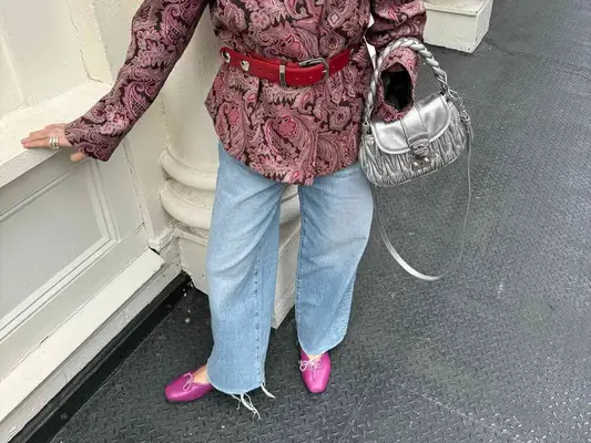 Close-up of April Lockhart wearing wide leg light wash jeans, pink flats, silver handbag, patterned blazer, and red belt