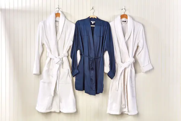 Three bathrobes, two white and one navy blue, on hangers set against a cream wall