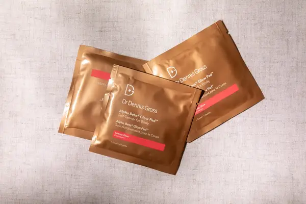 Three packets of Dr. Dennis Gross self-tanner wipes for body, arranged on a fabric surface