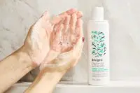 A person lathering Briogeo Destined For Density Caffeine + Biotin Density Shampoo in their hands next to the bottle